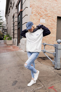 Person wearing a white sweatshirt with black sleeves and blue jeans on a city street.