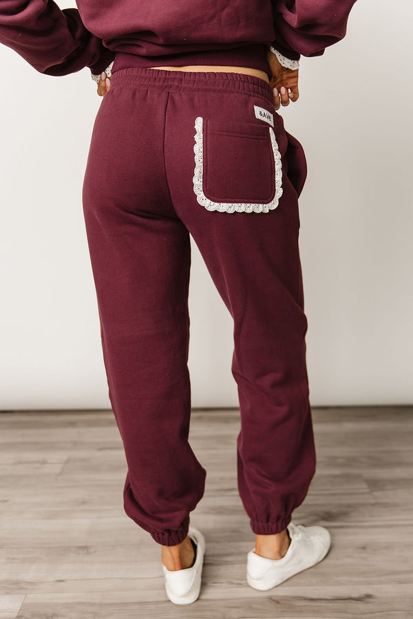 Maroon sweatpants with white lace trim on pockets worn by a person against a plain background